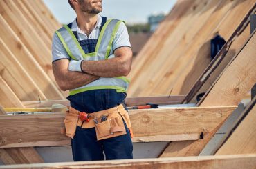 Cropped photo of a self-assured man wearing a tool-belt while standing cross-armed near construction in progress